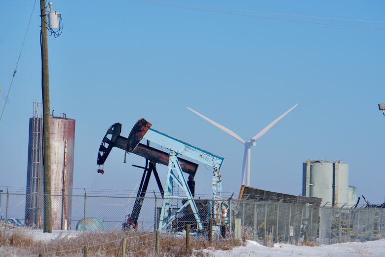 A wind turbine is pictured behind a pair of oil pumpjacks and a storage tank.