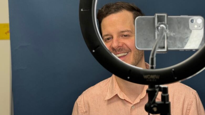 A smiling man is seen through a ring light. A camera is mounted in the centre and pointed at him. 