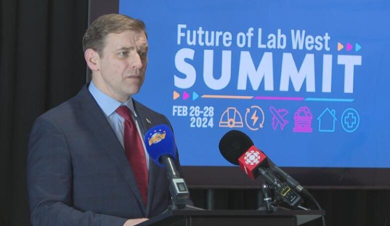 A man wearing a suit stands at a podium in front of a large screen. The screen shows the logo for the Future of Labrador West Summit.