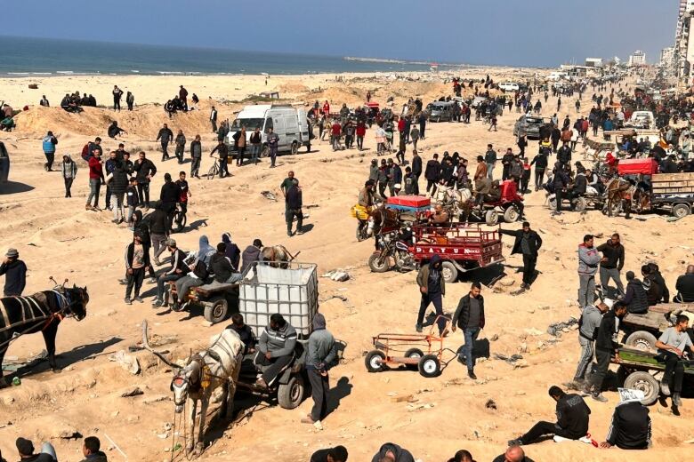 A large crowd of people waiting among carts, wagons and vehicles along a stretch of beach.