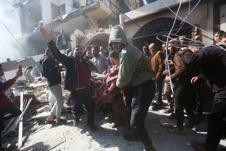 A group of people carry another person in a blanket away from the rubble of a building hit by an airstrike. 