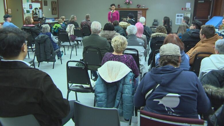 A group of people are seen sitting in chairs, facing a woman in front of them speaking at a microphone.