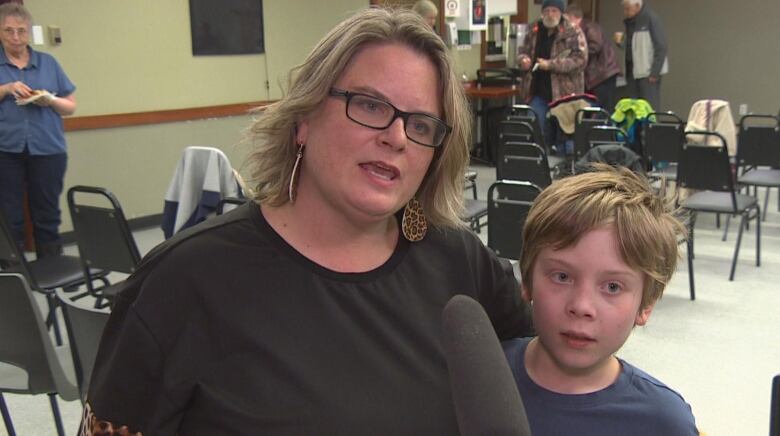 A woman is seen speaking at a microphone, while holding an arm around her younger son.