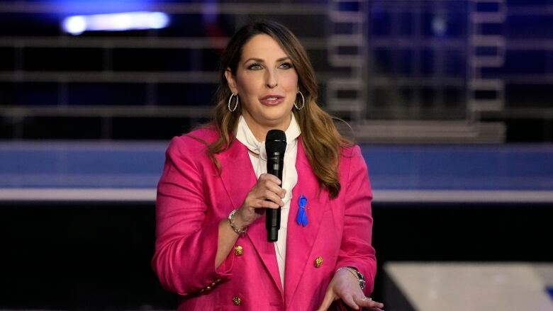 A woman in a blazer and a collared shirt is shown speaking while holding a microphone.