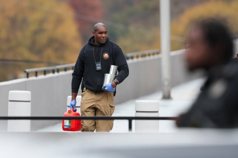 A dark-complected man wearing a hoodie holds metal cans in one arm, and a gasoline cannister in another.