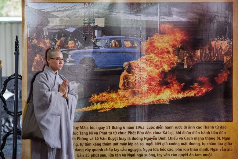 A bald, bespectacled person stands in front of a famous picture of a man set afire.