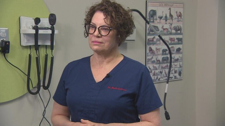 A woman wearing glasses standing in a doctor's office wearing medical scrubs.