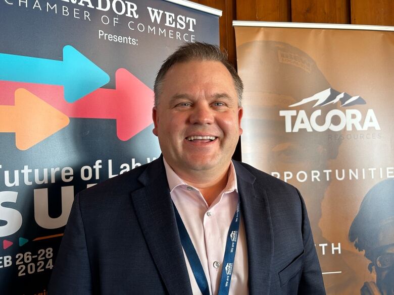 A smiling man wearing a suit stands in front of two banners. One is for Tacora, and the other is for the Future of Labrador West summit.