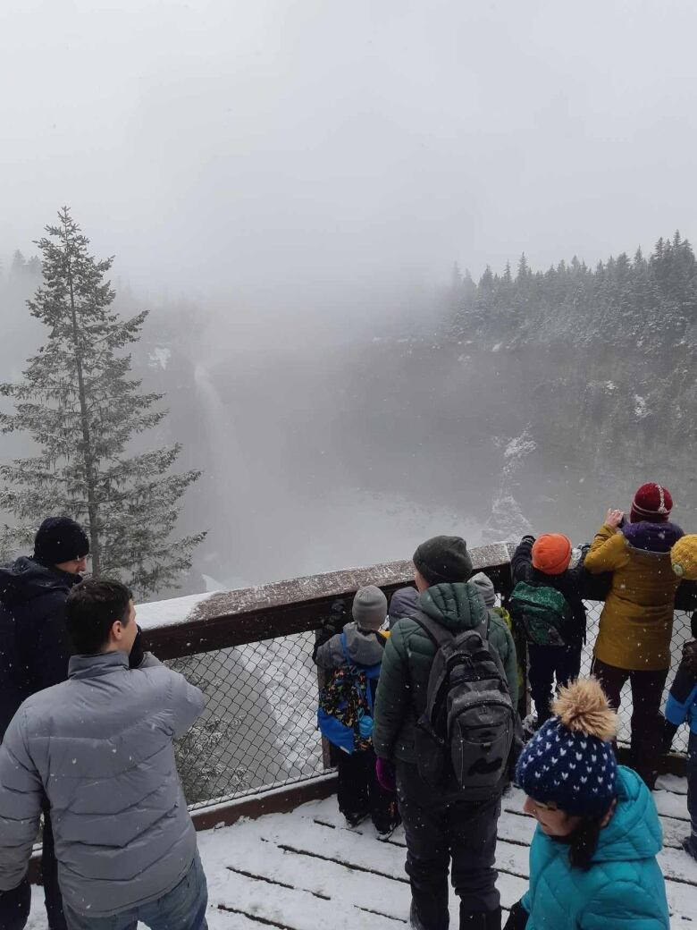 Group of people stand on a wooden viewing platform covered in snow looking at a winter waterfall.