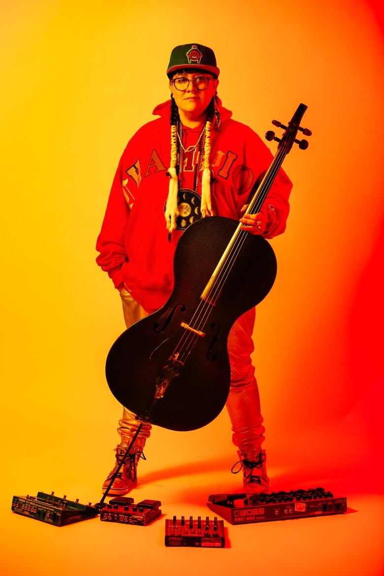 A woman, holding a black cello in her left hand, is wearing all orange. She is standing in front of an orange background.