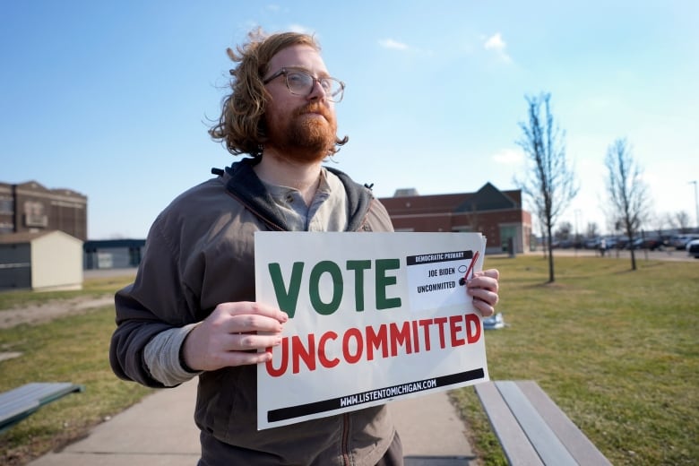 A man stands outside holding a white paper sign that reads 