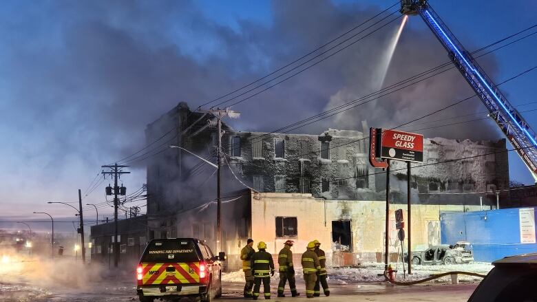 Firefighters spray water onto a burning building from a ladder truck. Thick smoke fills the sky above the three-storey building.