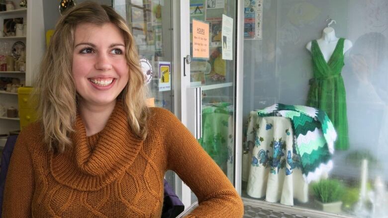 A smiling young woman standing near a window with a dress in the background.