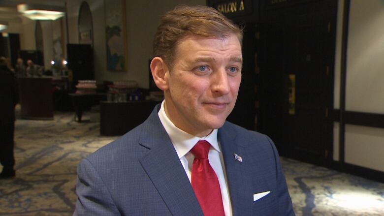 A man wearing a blue suit and a red tie stands in a hallway.