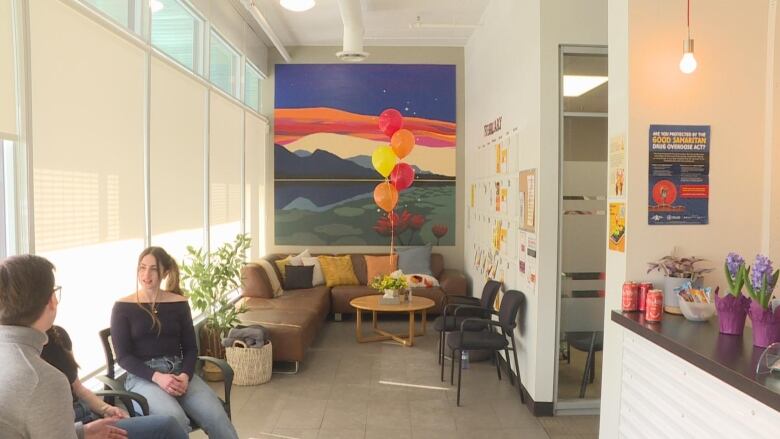 a room with a couch and chairs. there is a reception-style desk decorated with flowers. two people sit in chairs by a window and chat.