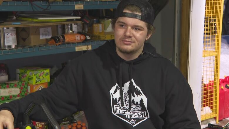 A man wearing a sweater with the Cain's Quest logo on it sits on a snowmobile in a warehouse.