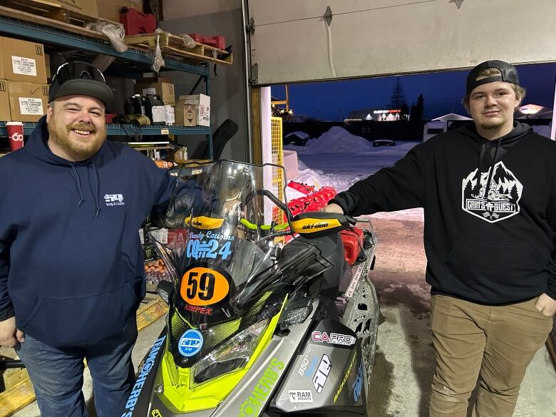 Two smiling men stand on either side of a snowmobile.