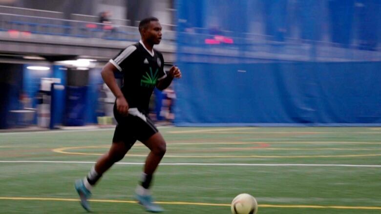 A soccer player in an indoor field chases a ball. He is wearing a black, white, and green uniform and the background is blurred. 