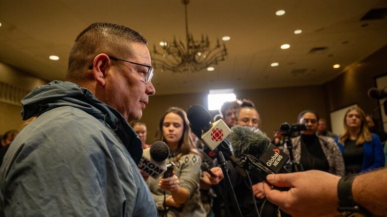 A man in a gray jacket speaks to media with microphones out