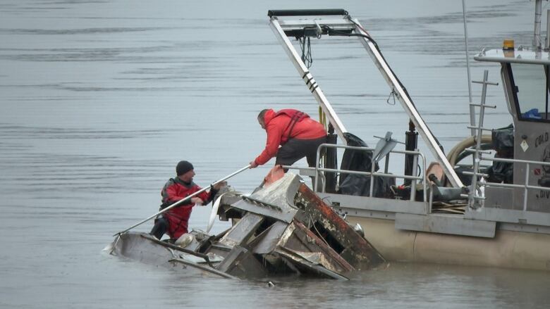 A man in a dry suit is on a mostly-submerged boat, which is brown and nasty from years of neglect in the the river. A second man is on another boat that's still perfectly functional. He's assisting with a pole.