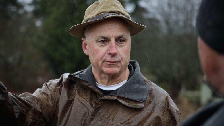 A man wearing a crumpled brown brimmed hat and wet brown rain jacket speaks to another man who is out of focus and barely in the frame.