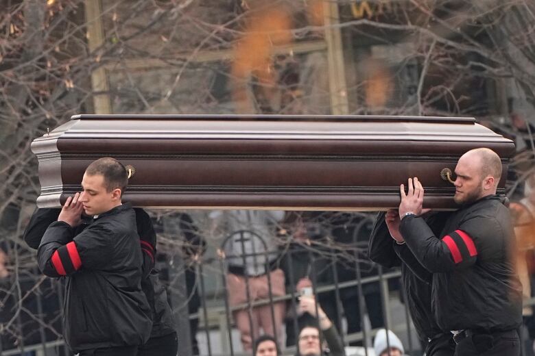 From a distance, four men are shown lifting a coffin on their shoulders. The faces of two men are visible. 