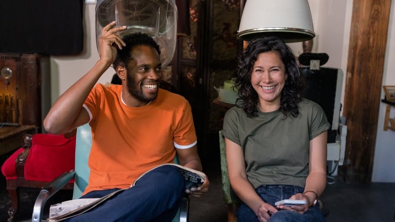 A man and a woman sit under old fashioned hairy dyers in a salon. They are both smiling.