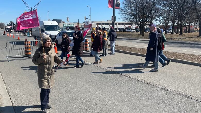 A woman waves a flag on a gated off road. There is a picket line in the background. It's a blue, sunny, early spring day