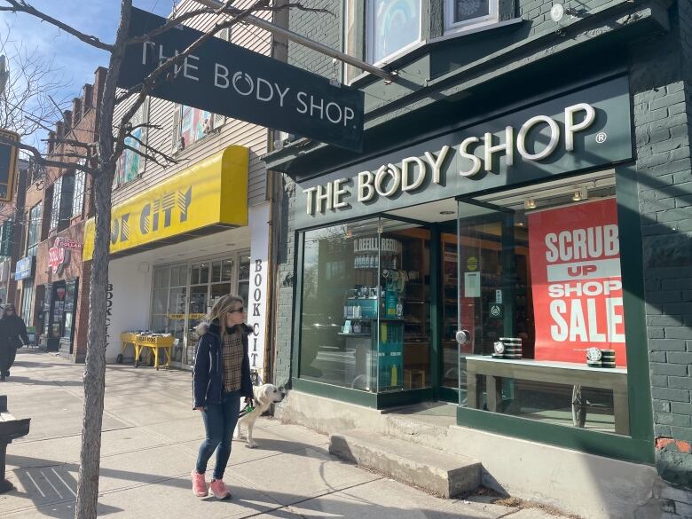 A woman and her dog walk by a storefront with a sign reading The Body Shop.