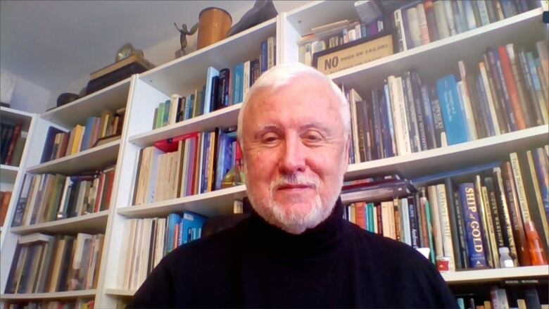 A man sits in front of a white bookcase filled with books.