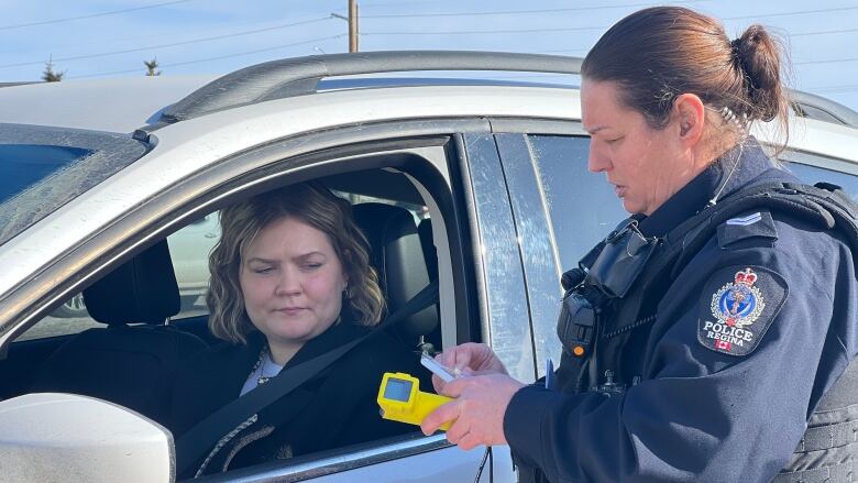 A police officer stands outside a vehicle holding a yellow device, while the driver of a vehicle sits inside the car.