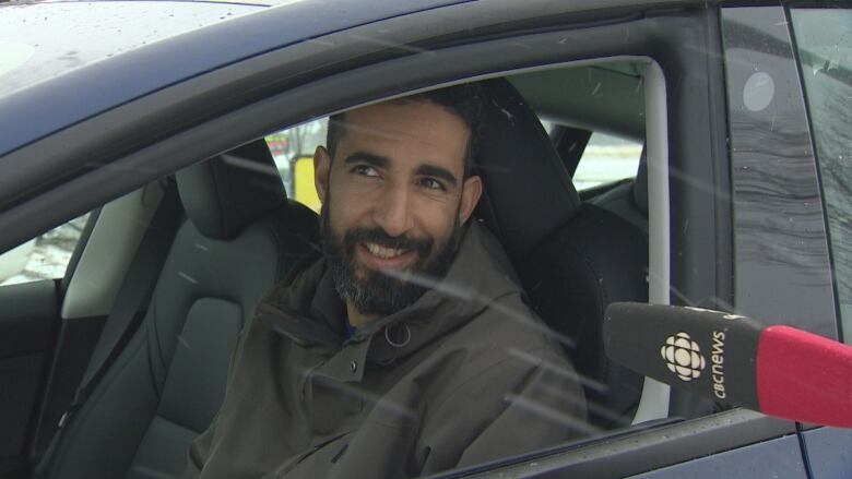 A man with dark hair and a beard sitting inside of a blue electric vehicle on a snowy day.