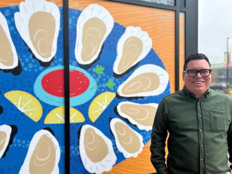 A person wearing thick, black glasses stands smiling in front of an art piece. 