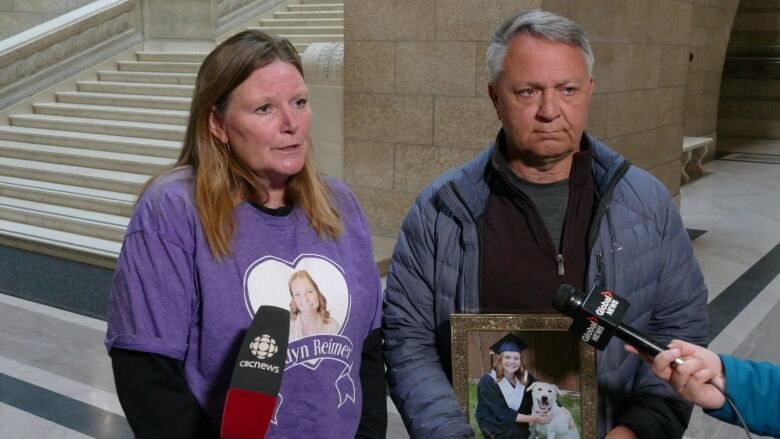 A woman in a purple t-shirt and a man in a blue jacket speak with members of the media.