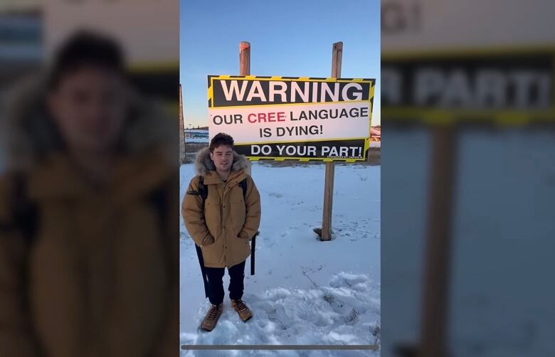 A man wearing a beige jacket is standing in front of a sign that says 'Warning our Cree language is dying, do your part'