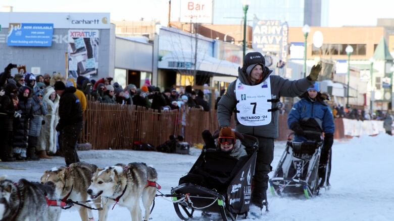 A person in heavy winter outerwear waves to people off camera as they stand on the back of a sled pulled by dogs.