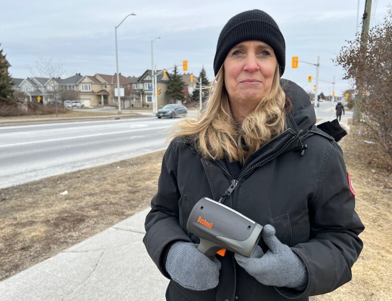 A woman holds a speed radar gun alongside a road.