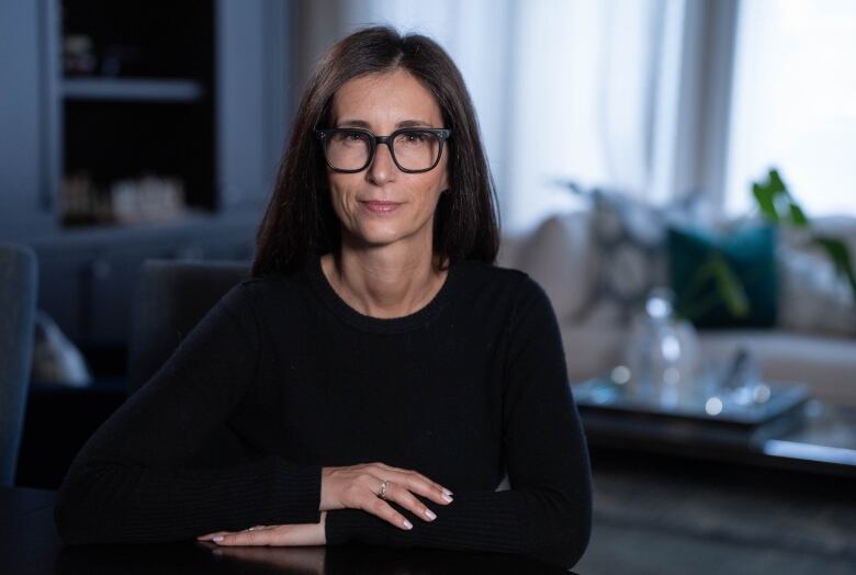 A woman with long hair, seated wearing a black top and black glasses. 