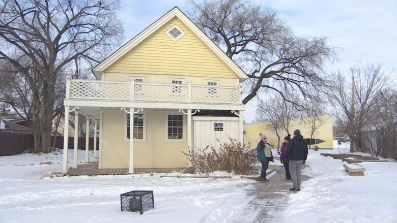 Two women speak with a man in a toque outside a historic yellow two-storey house surrounded by snow on the ground.