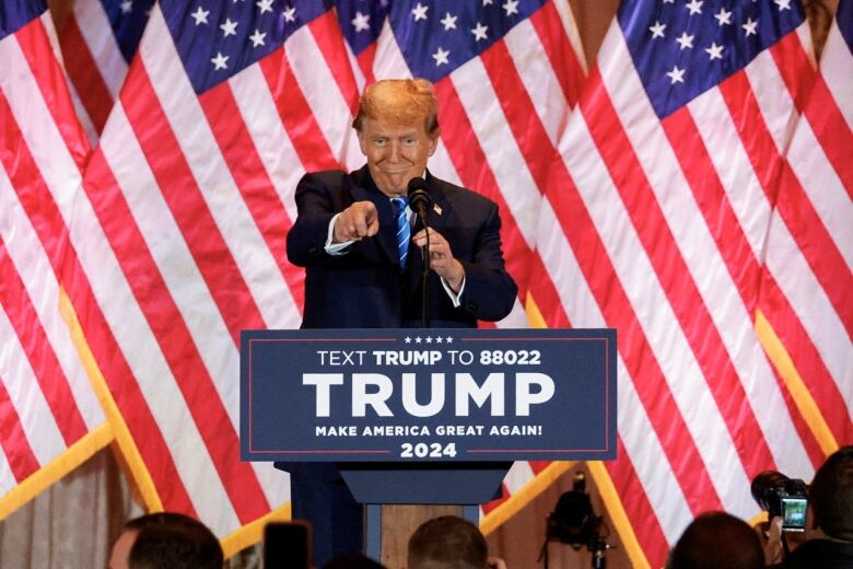 Donald Trump stands at a podium, in front of a row of U.S. flags, with a smirk on his face and pointing his finger at the audience in front of him.