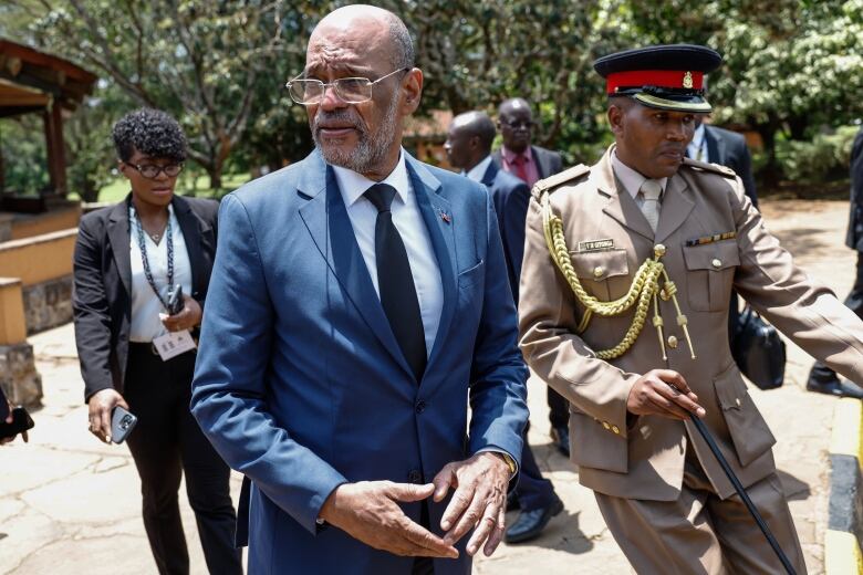 A bespectacled, bearded balding man in a suit and tie walks outside beside a man in a military uniform. Other people, men and women, are shown in the background.