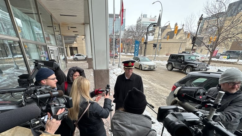 A man in a police uniform stands outside a building surrounded by people holding microphones and cameras.