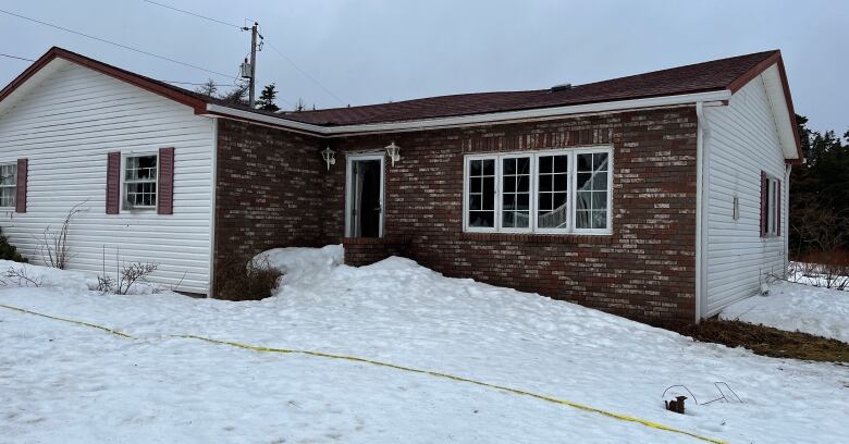 A bungalow with white and brick siding sits behind a snow-covered yard. Yellow police tape can be seen on the ground.