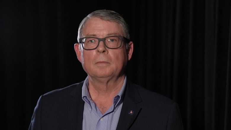 A man in a dark blazer wearing glasses looks toward the camera.