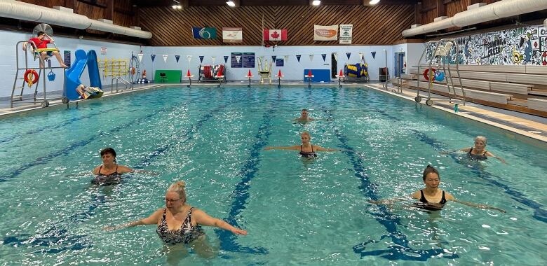 Six aquafit participants stand in the pool with both arms outstretched. 