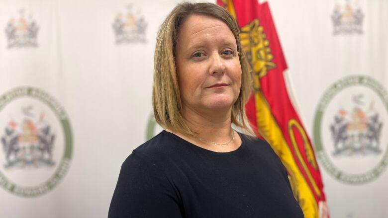 Jenn Redmond stands against a backdrop of flags. She is wearing a dark blue top and looks directly in to the camera.