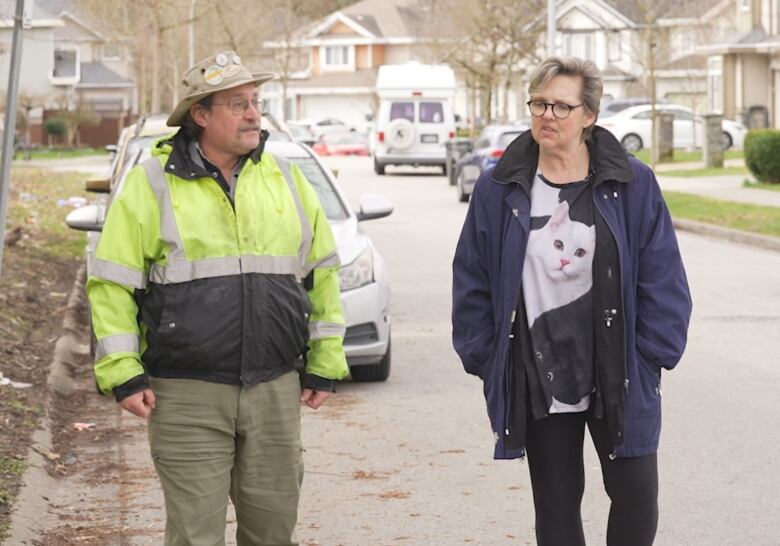 A man in a hat and hi-viz vest and a woman in a shirt with a cat image walk down a street