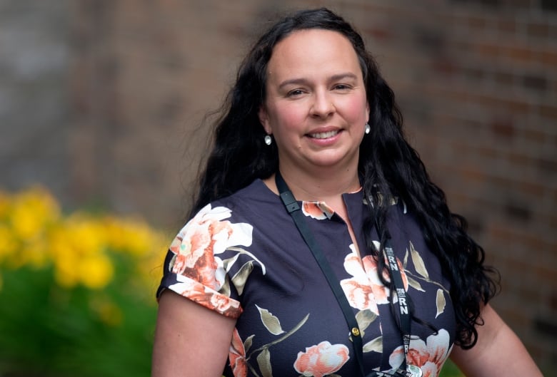 A woman stands with yellow flowers and a brick wall in the background.