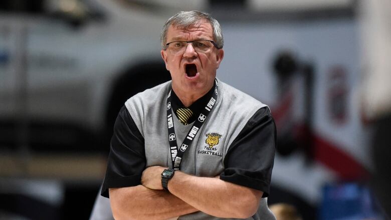 A man in a grey vest, and black-t-shirt, stands on the side of a basketball court. His mouth is open as if in mid yell. 