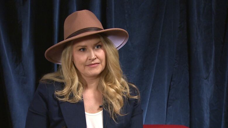 A woman with long blonde hair wearing a sunhat sits in front of a blue curtain.